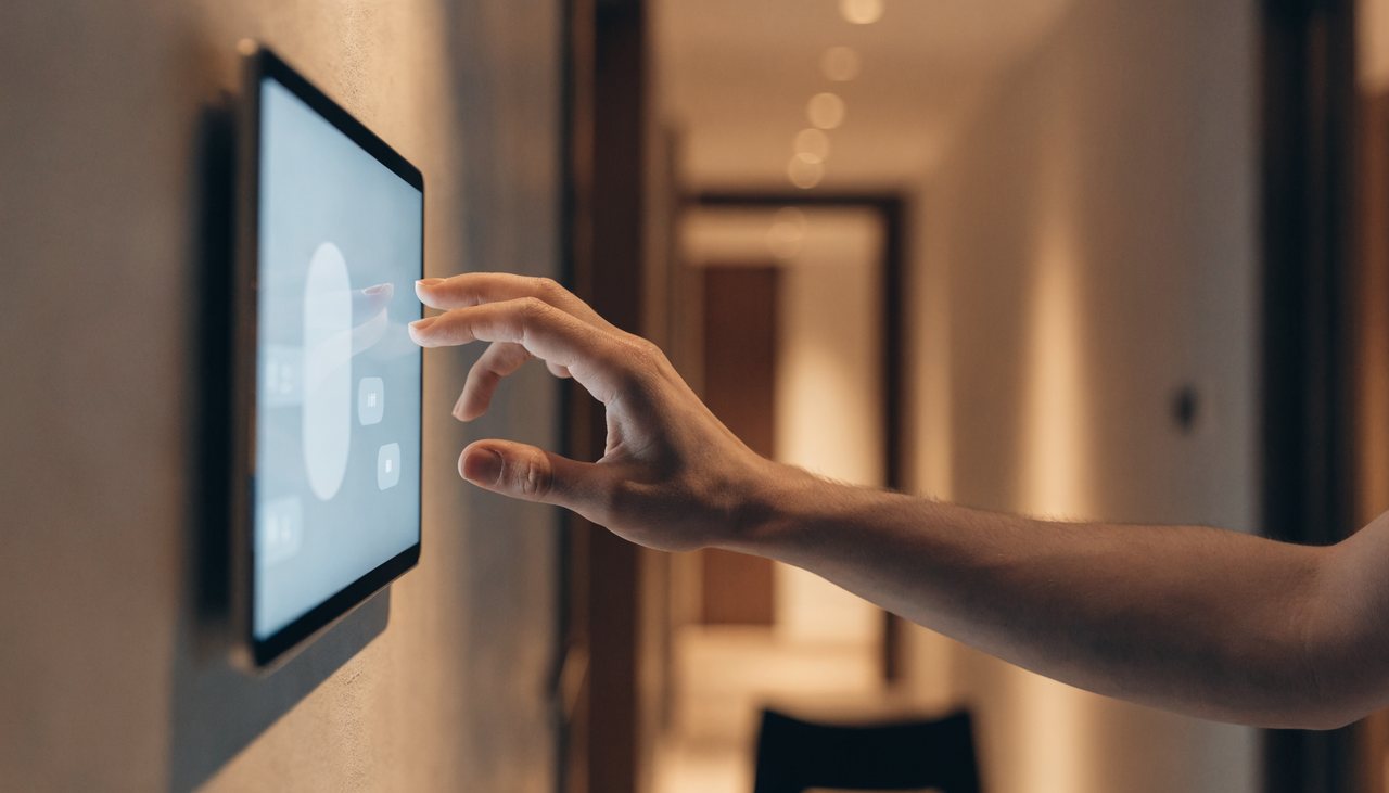 A hand tapping a wall-mounted smart-home control tablet showing rooms and scenes